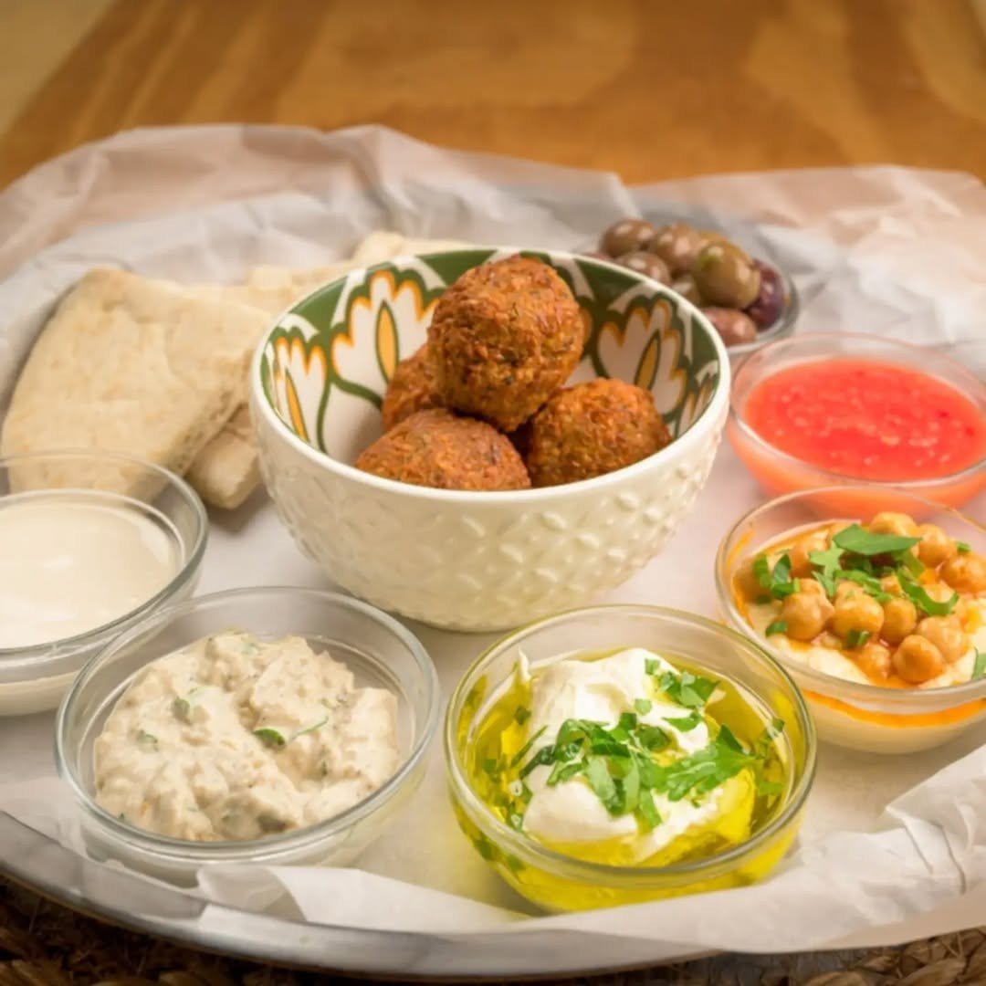 Tabuleiro com uma taça de falafel, pão pita e taças de pastas cremosas, grão-de-bico e molho vermelho