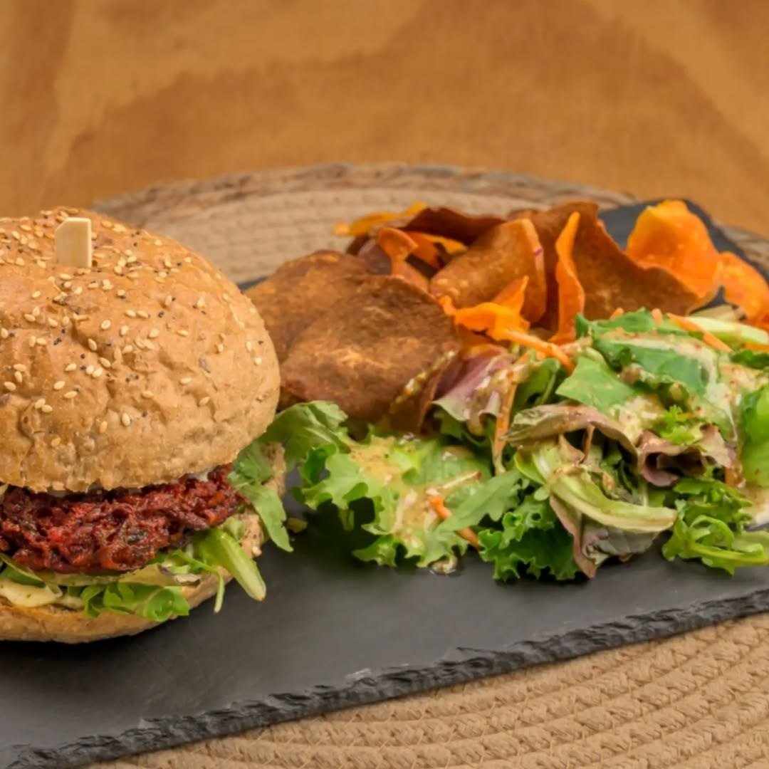 Hambúrguer vegetariano em pão com sésamo, ao lado de salada verde e chips de batata-doce, numa ardósia