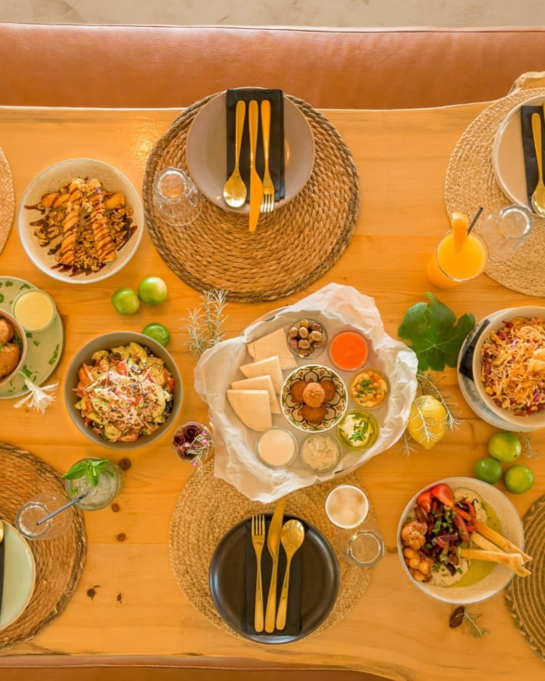 Vista de cima de uma mesa de madeira com várias taças de saladas, pastas, pão pita e limas, à volta de individuais de palha